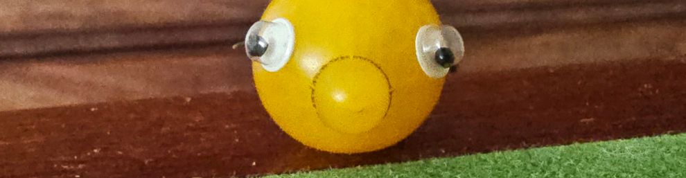 A yellow tomato that has grown in a strange way with a blob at the front, which makes it look like a nose. It has eyes stuck on it and two of the green stalk fronds stick out behind it but because it’s shown from the front they look like green ears. It sits on a green baize table with a line of veneered wood at the side. Behind it is a big brown chest of drawers.