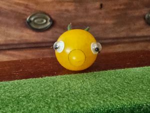 A yellow tomato that has grown in a strange way with a blob at the front, which makes it look like a nose. It has eyes stuck on it and two of the green stalk fronds stick out behind it but because it’s shown from the front they look like green ears. It sits on a green baize table with a line of veneered wood at the side. Behind it is a big brown chest of drawers.