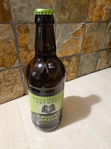 PIcture of a bottle of beer on a light coloured wooden surface against a reddy-brown and grey tiled wall. The bottle is brown and crown top is lime green. The label reads ‘Roughacre Brewery’ on a light green background. Below is a white stripe with a black and white graphic of an opened book.Below reads ‘Foreword Festival’ which is also the festival logo, done in a font that’s a little like graffiti tags. Below this in smaller letters, the beer is described, ‘Golden pale ale, 3.6% ABV’ 
