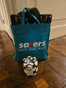 a turquoise shopping bag with red and white writing on it srabding against a white panelled wall on a chequered wooden floor. in front of t,he bag is a loo roll wrapped in jazzy black and white paper. this design is white circles. in the bag you can make out a dark coloured canvas and metallic object which is clearly folded.