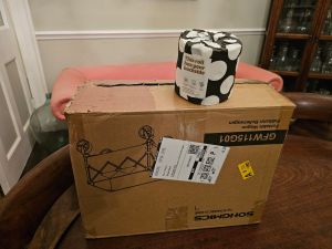 A brown cardboard box with a loo roll wrapped in jazzy black and white paper sitting on top of it to give a feel of size. It sits in front of a red chesterfield which is set against a beige wall with a white dado rail.