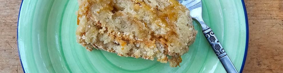swiss roll style cake, plain cream/off white plain sponge coloured with yellow icing and a fork beside it on a green plate, with a thin blue rim, placed on a light coloured wooden table.