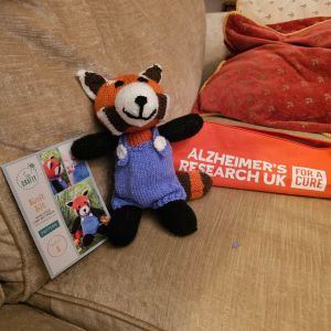 Picture of a knitted bear in brown, black and white wool with blue knitted dungarees. It’s on a beige couch with an orange knitting needle case bearing the legend Alzheirmer’s Research UK and behind that a brown/red cushion. Next to the bear is the pattern with a picture of how it’s supposed to look on the front. Mine isn’t too far off.