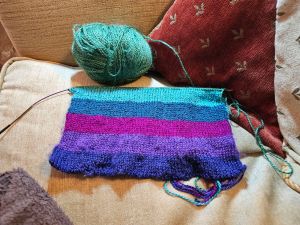 Picture of a knitted scarf in progress on a single loop needle in blue, mauve, purple, teal and turquoise wool with a ball of green. It’s all sitting on a beige sofa with a red cushion with beige fleur de lys on it behind.