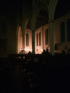 Picture of a darkened church with a distant altar, candles lit and someone reading from a lectern