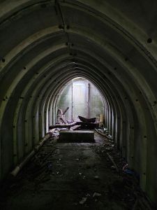 Looking into a dark narrow vaulted building with light falling from a skylight at the far end against a concrete back wall. Debris is also present, a concrete block, wood, cans and the hopper of an old wheelbarrow