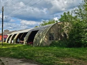 Old aircraft hanger with cloudy and blue sky and green verge in front plus trees to the right and a telegraph pole to the left